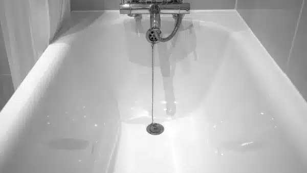 A black and white image of an empty bathtub features a central drain, ready for any drain cleaning tasks. A chain is attached to the plug, and a faucet is mounted above.