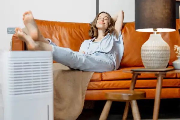 A woman unwinds on a brown sofa, legs up and eyes closed, by a dehumidifier needing humidifier repair, under the gentle glow of a lamp.
