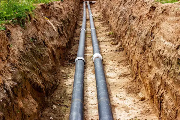 A trench filled with two parallel black pipes laid underground, surrounded by soil, is an essential step in the sewer line installation process.